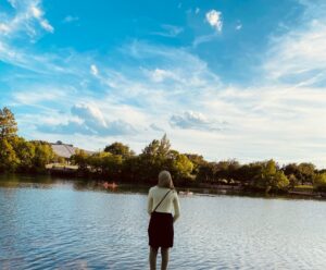 A girl standing in front of a lake with trees in the background.