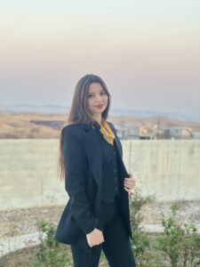 Shifaa has long dark hair. She is wearing a black pantsuit.