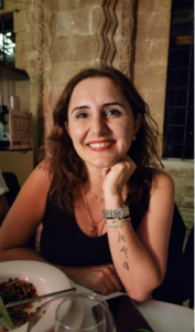Figen is a woman with brown hair. She is leaning her face on her hand as she smiles.