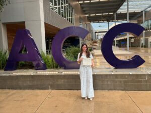 A girl in pinstripe clothes stands in front of the ACC sign holding an award certificate.
