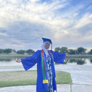 A woman in graduation robe and cap proudly spreads her arms.