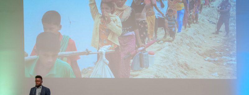Phillip A. Townsend speaking in front of a slide featuring women and children immigrating overseen by a guard